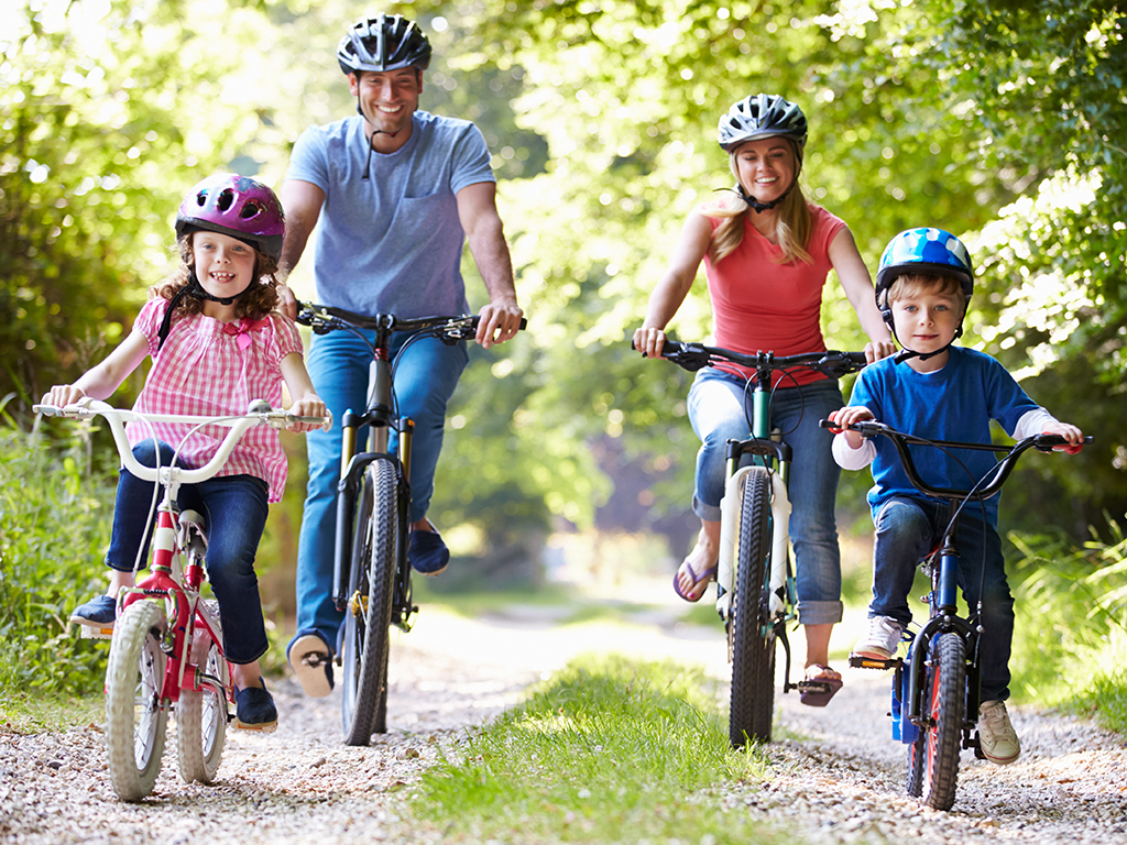 A family of four rides bicycles on a gravel path through a wooded area. All are wearing helmets. The parents and two children are smiling and appear to be enjoying the outdoor activity.